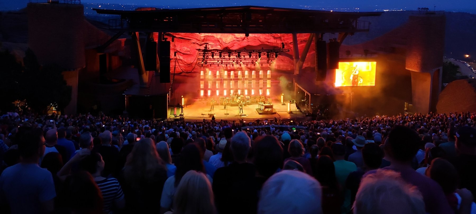 Red Rocks Amphitheatre at dusk, red-lit rock formation behind the stage, crowd silhouetted in the foreground